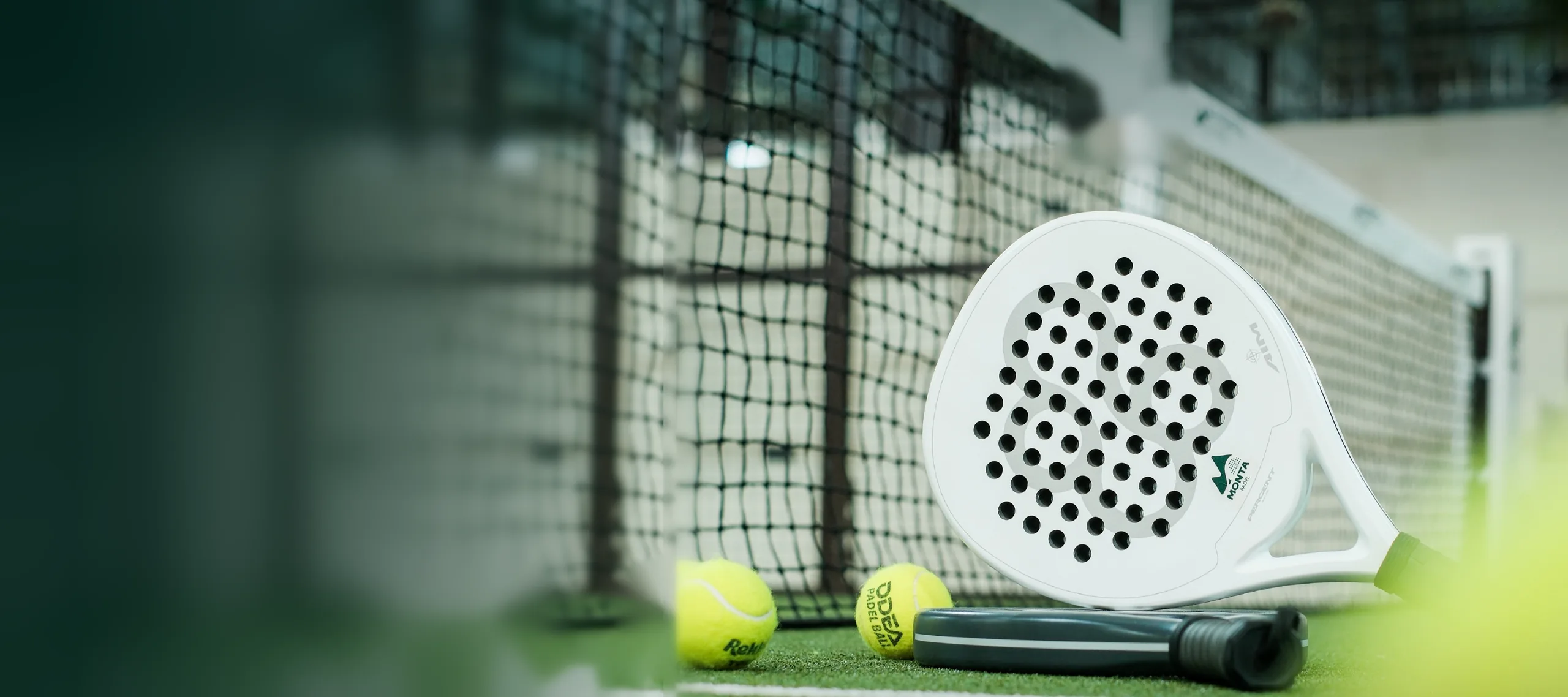 A white Monta padel racket and two balls resting against the net on the green court