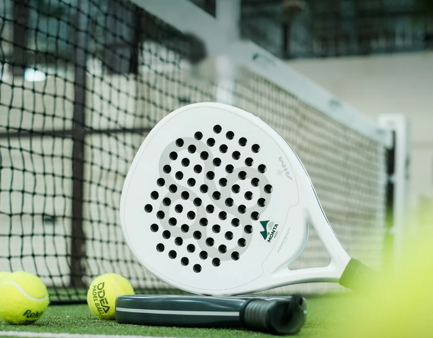 A white Monta padel racket and two balls resting against the net on the green court