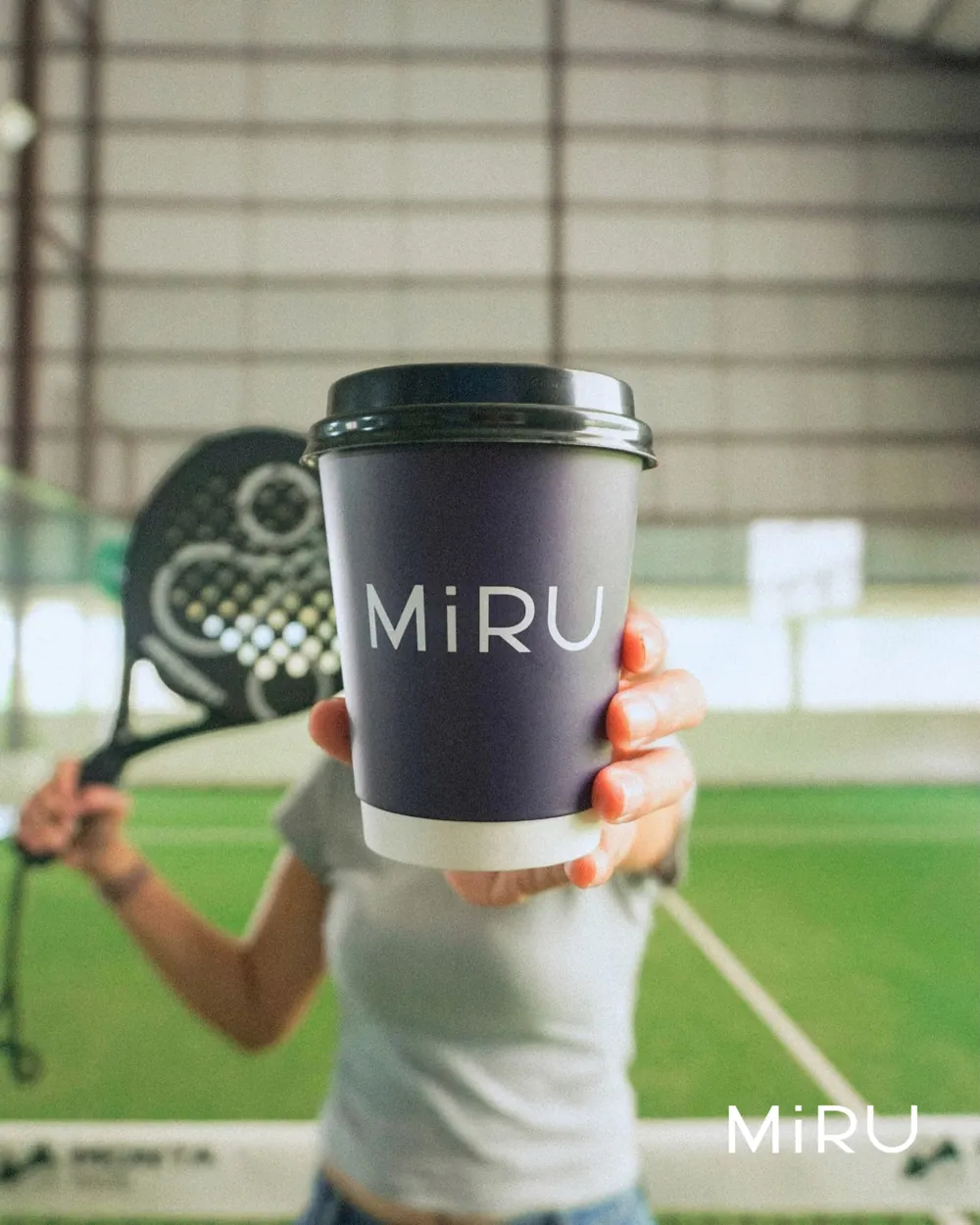 A Miru takeaway cup held up on court, a racket behind it